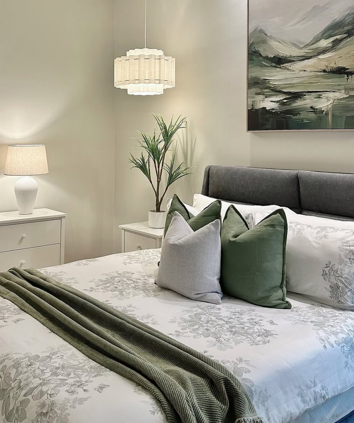 A decorated bed with grey and green pillows, white floral linens, and a textured throw blanket.