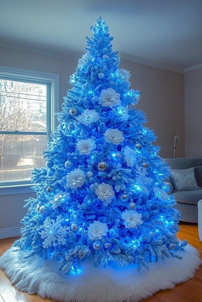A bright blue illuminated Christmas tree decorated with white flowers and silver ornaments on a white skirt.