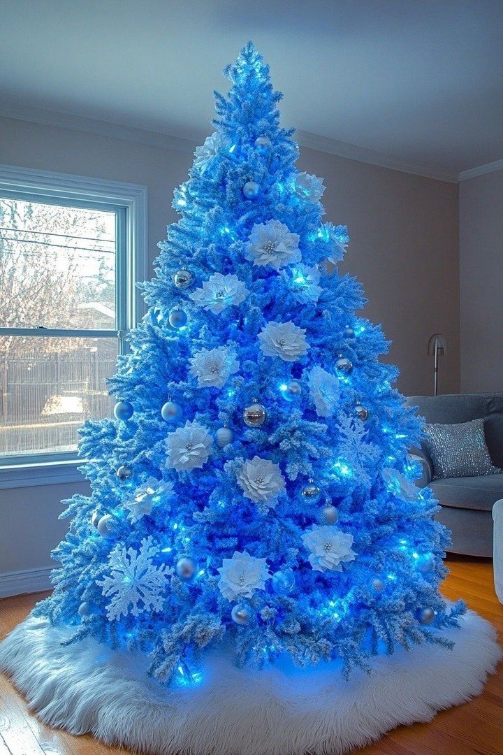 A bright blue illuminated Christmas tree decorated with white flowers and silver ornaments on a white skirt.