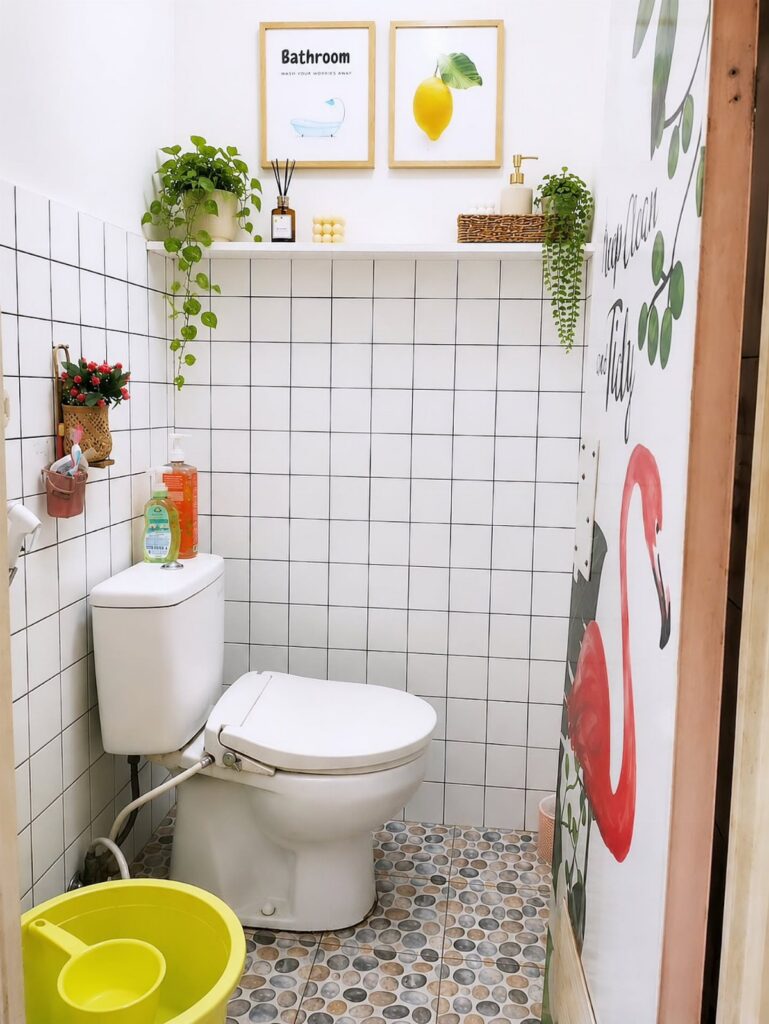 A small white tiled bathroom with a toilet, plants on a shelf, and flamingo wall art.