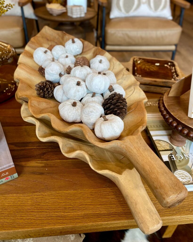 White decorative pumpkins and pinecones inside a long, wavy wooden dough bowl on a table.