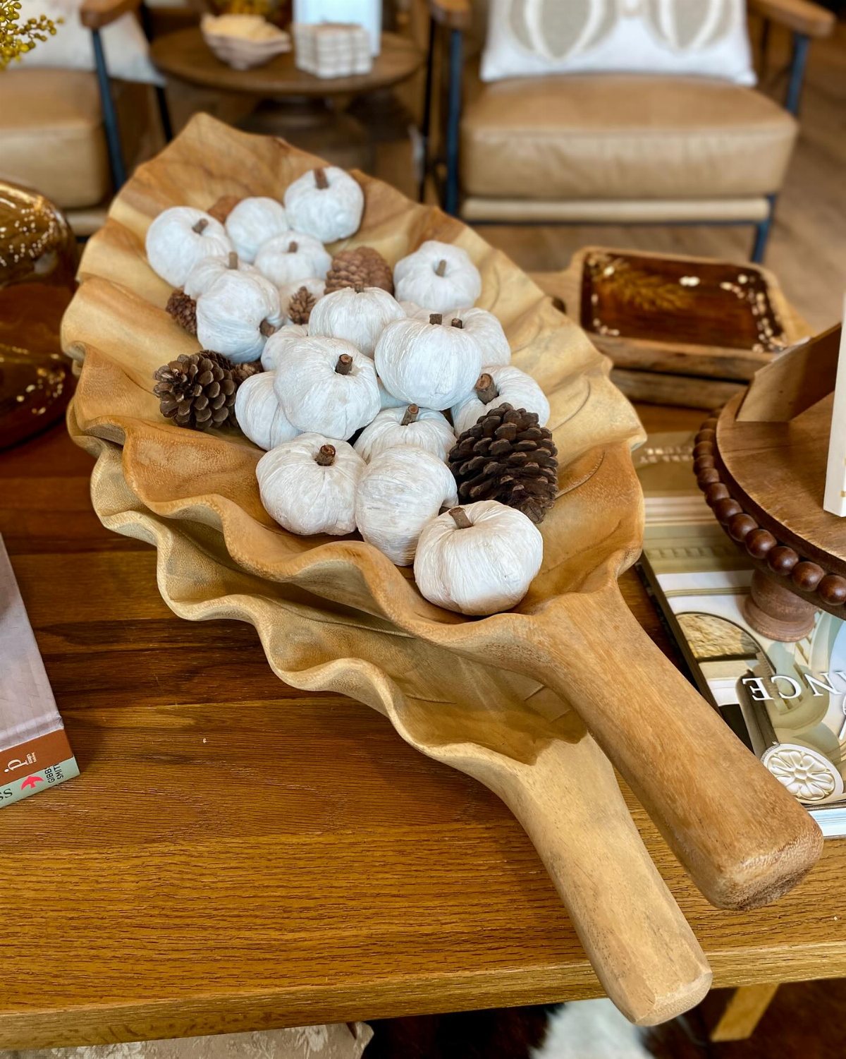 White decorative pumpkins and pinecones inside a long, wavy wooden dough bowl on a table.