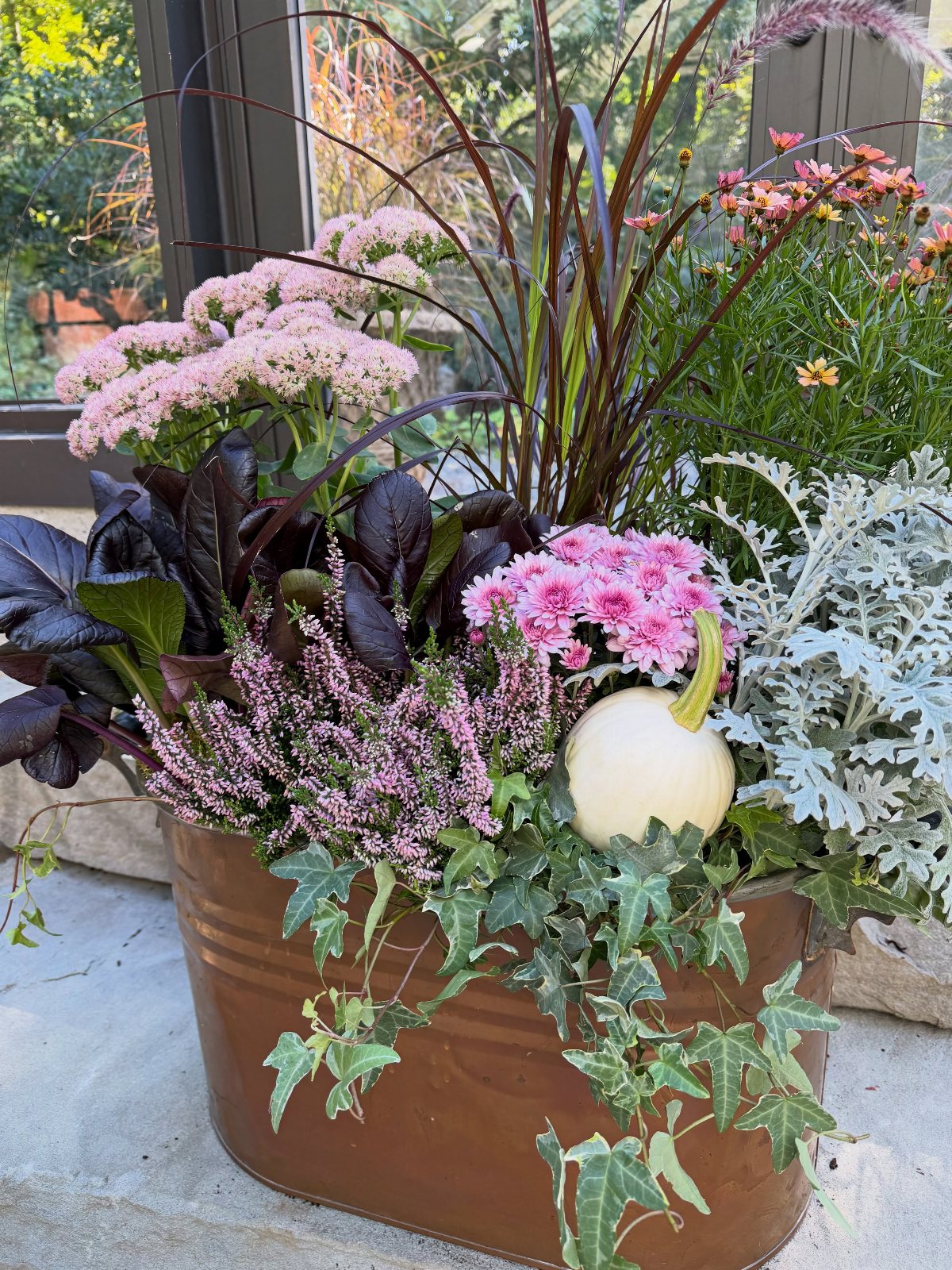 A metal planter filled with pink flowers, purple heather, and a small white pumpkin.