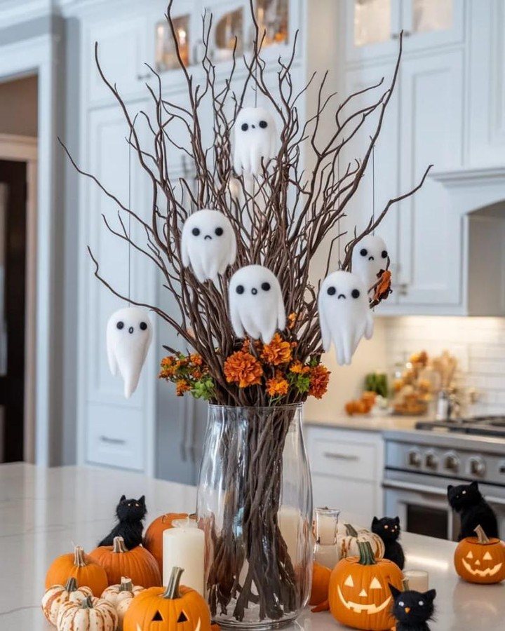 White ghost decorations hanging from branches in a vase surrounded by pumpkins on a counter.