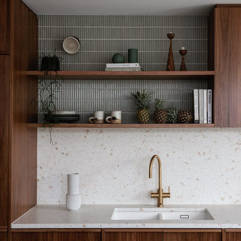 Wooden open kitchen shelves styled with ceramics, books, and pineapples against a grey tiled backsplash.