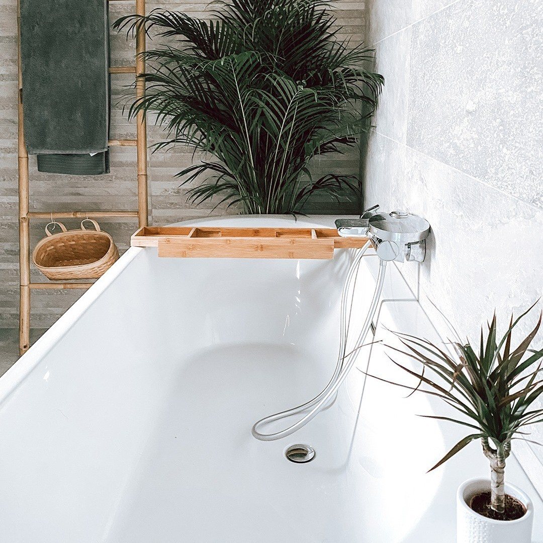 White bathtub with a wooden tray, surrounded by lush green tropical plants and bamboo decor.