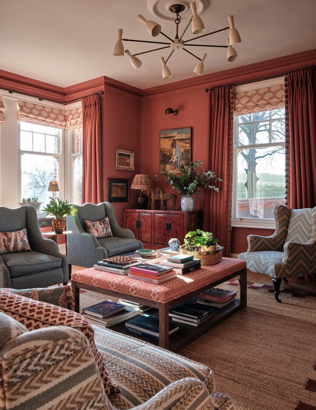 Terracotta-red living room featuring patterned armchairs, a large ottoman, and a modern chandelier.