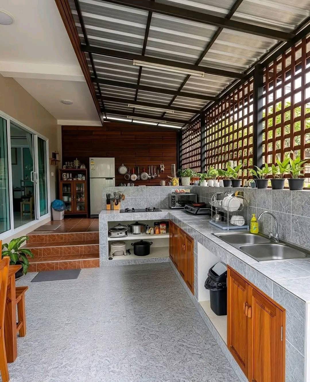 Outdoor kitchen with grey tiled counters, wooden cabinets, and potted plants under a metal roof.