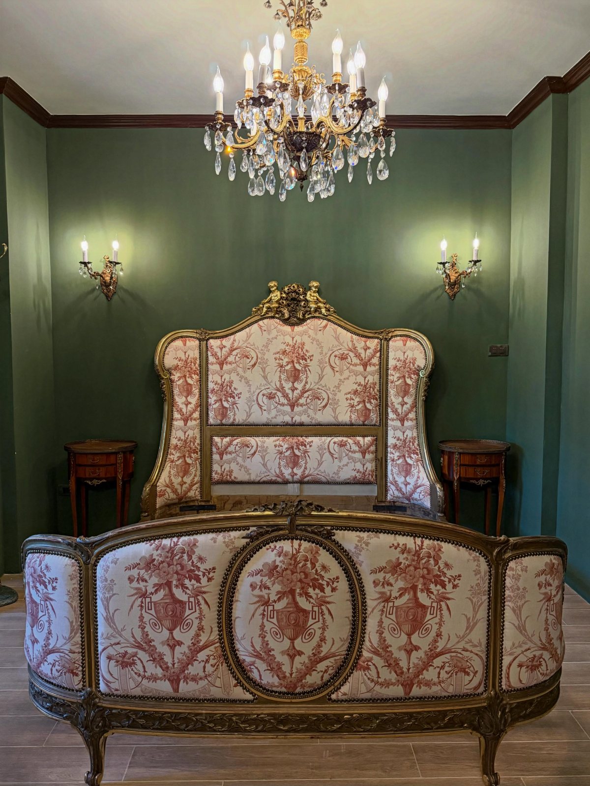 Ornate Victorian bed with red damask upholstery against dark green walls with a crystal chandelier.