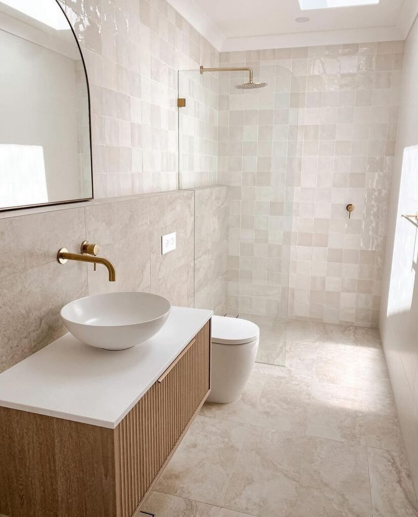 Modern bathroom with beige tiles, gold fixtures, a wooden vanity, and a walk-in shower.