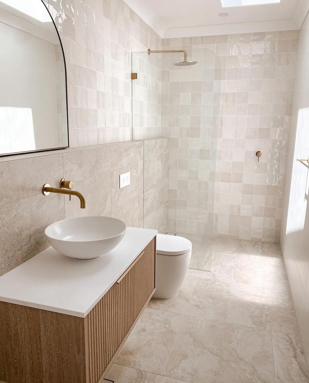 Modern bathroom with beige tiles, gold fixtures, a wooden vanity, and a walk-in shower.