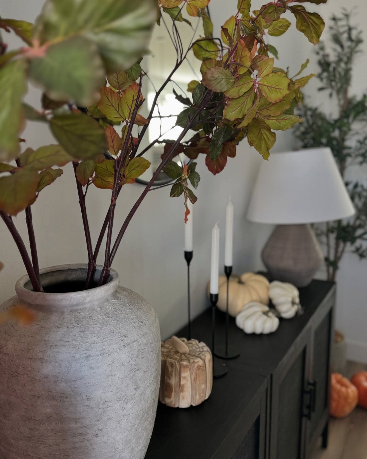 Autumn branches in a grey vase sit beside white pumpkins and candles on a black console table.